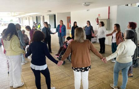 Cerca de duas dezenas de pessoas formam uma roda (círculo) aparentando estarem a dançar. Estão quase todas de mãos dadas. No centro da roda está uma pessoa em cadeira de rodas.