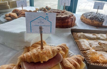 Plano aproximado de vários doces e aperitivos (em cima de uma mesa). Junto dos produtos comestíveis está um monte de guardanapos. Em primeiro plano surge um croissant com um pequeno palito no qual foi colado um papel com a imagem de uma casa e o texto: Café na Casa. Dia da Família das Unidades Residenciais. 18 de maio.