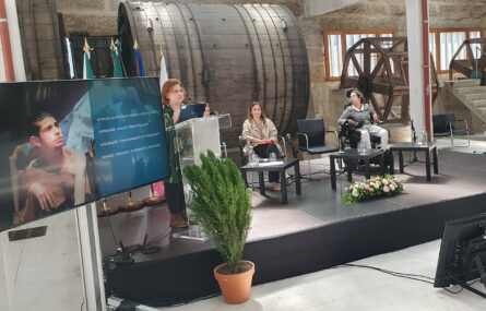 Fotografia de pequeno palco no qual estão quatro pessoas. Uma das pessoas fala junto do púlpito. Do lado esquerdo da imagem projeção em ecrã. Ao fundo vários equipamentos, com aspeto antigo, de aparente utilização industrial.
