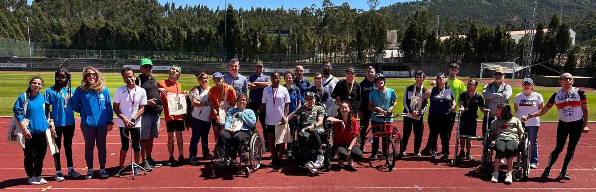 Fotografia de um grupo com 29 pessoas, lado a lado, em pista de atletismo. Ao fundo vê-se o relvado do estádio e em paisagem imensas árvores e um monte.