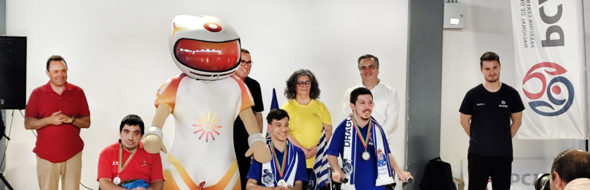 Grupo de oito pessoas alinhadas junto de um pódio improvisado em pavilhão desportivo. Além dos três primeiros classificados, atrás deles surgem ainda outras pessoas e uma mascote. Em primeiro plano, de costas, são parcialmente visíveis duas pessoas a fotografar o momento.