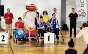 Grupo de oito pessoas alinhadas junto de um pódio improvisado em pavilhão desportivo. Além dos três primeiros classificados, atrás deles surgem ainda outras pessoas e uma mascote. Em primeiro plano, de costas, são parcialmente visíveis duas pessoas a fotografar o momento.