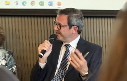 Pessoa sentada, a falar, com um microfone na mão. Atrás dessa pessoa vê-se um pouco de uma projeção em tela.