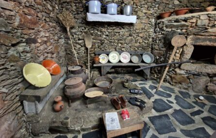 Fotografia de espaço interior com chão e paredes em pedra. No local estão espalhados inúmeros utensílios antigos. Vê-se, parcialmente, um forno.