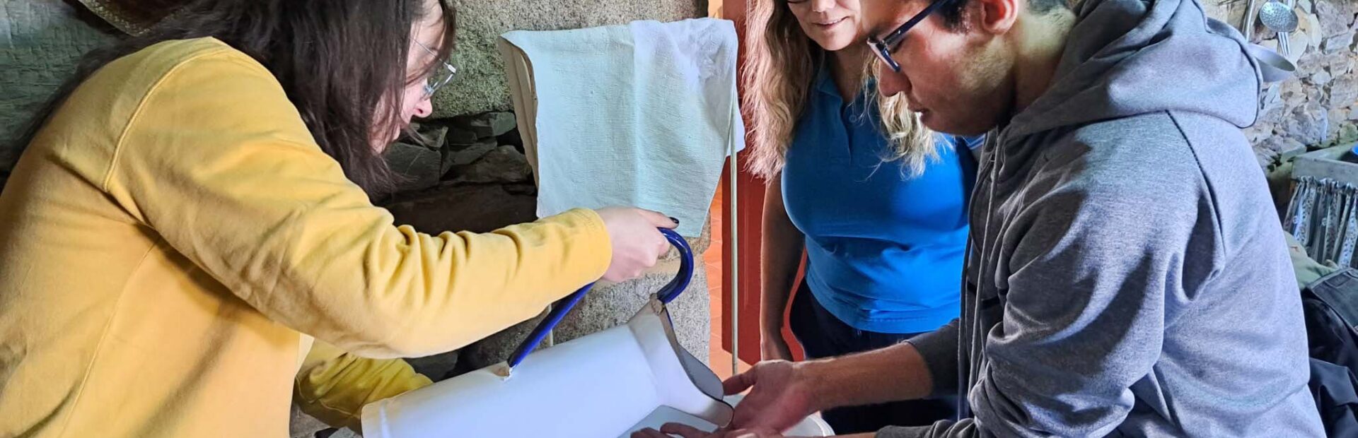 Três pessoas em espaço interior com paredes em pedra. Uma das pessoas usa um jarro antigo para despejar água nas mãos de outra.