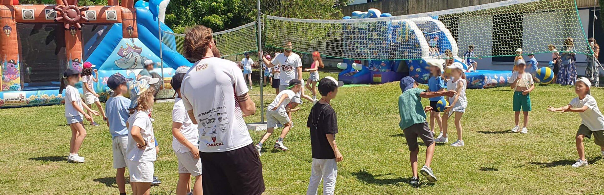 Várias dezenas de crianças em atividade física em jardim relvado exterior. Ao fundo da imagem um insuflável gigante, algumas árvores e um edifício. Os rostos visíveis das crianças foram digitalmente desfocados.