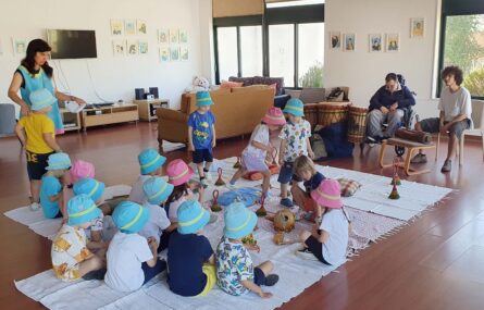 Quase duas dezenas de crianças em espaço interior. Estão acompanhadas por três adultos. Junto de dois dos adultos estão vários instrumentos musicais. Os rostos visíveis das crianças foram digitalmente desfocados.