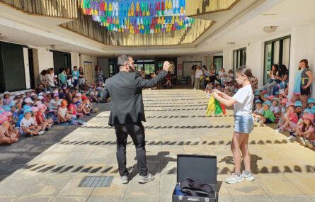 Perto de uma centena de crianças sentadas em espaço exterior. Ao centro da imagem um adulto aparenta fazer magia. Junto dele está uma criança a colaborar. Os rostos visíveis das crianças foram digitalmente desfocados.