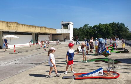 Várias dezenas de crianças em espaço exterior amplo. Estão a praticar várias atividades desportivas. Os rostos visíveis das crianças foram digitalmente desfocados. Algumas árvores ao fundo da imagem.