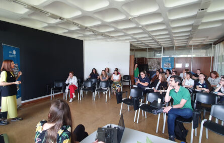 Sala interior com várias dezenas de cadeiras e pessoas sentadas. Do lado esquerdo da imagem uma pessoa gesticula e aparenta estar a falar. Em primeiro plano, de costas, vista parcial de uma pessoa a utilizar um computador portátil.