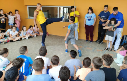 Várias dezenas de crianças sentadas no chão e em pé, em roda semi-circular. Estão a assistir a uma demonstração de Capoeira (realizada por uma pessoa adulta acompanhada de uma criança). Outros adultos e crianças tocam alguns instrumentos. Os rostos visíveis das crianças foram digitalmente desfocados.