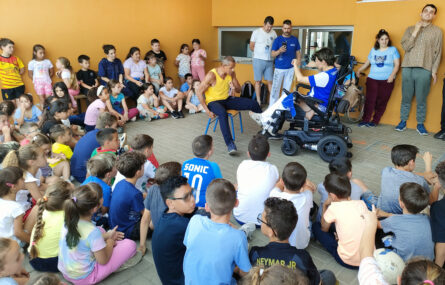 Várias dezenas de crianças sentadas no chão e em pé, em roda semi-circular. Estão a assistir a uma demonstração de Capoeira (realizada por duas pessoas adultas, sendo que uma delas está sentada numa cadeira e outra utiliza cadeira de rodas eletrónica). Outros adultos e crianças assistem de pé encostados a uma parede. Os rostos visíveis das crianças foram digitalmente desfocados.