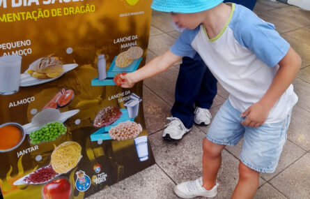 Uma criança aparenta estar a colar algo num cartaz gigante (com imagens de vários alimentos e bebidas). O rosto da criança foi digitalmente desfocado.
