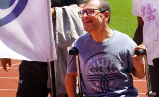 Plano aproximado de uma pessoa sorridente. Ao lado, parcialmente visível, bandeira da PCAND. Atrás são parcialmente visíveis duas pessoas e parte de uma pista e relvado.