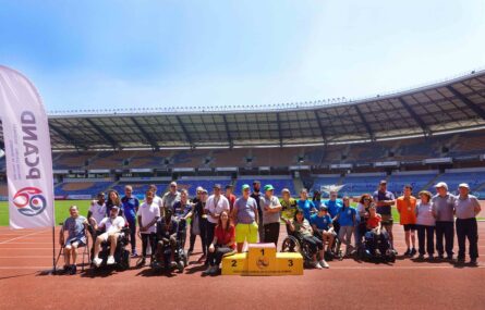 Fotografia de grupo, com 32 pessoas, na pista de corridas de um estádio. Ao fundo as bancadas e o céu azul. Em plano intermédio parte do relvado do estádio. Do lado esquerdo da imagem, junto do grupo, uma enorme bandeira com o logótipo da PCAND.