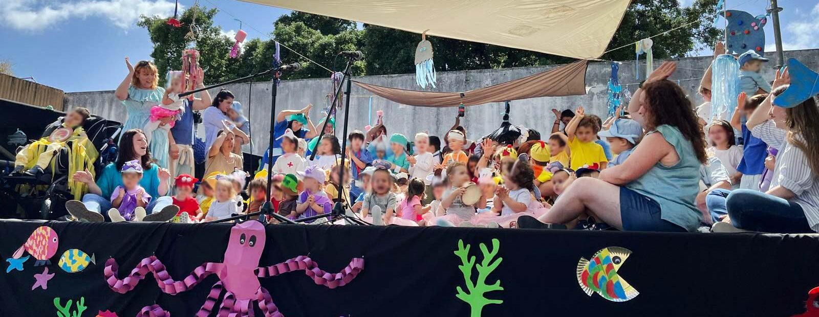 Palco com várias dezenas de crianças (e alguns adultos). Em primeiro plano, na parte frontal do palco, algumas imagens estilizadas com referências marítimas. Os rostos visíveis das crianças foram digitalmente desfocados.