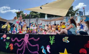 Palco com várias dezenas de crianças (e alguns adultos). Em primeiro plano, na parte frontal do palco, algumas imagens estilizadas com referências marítimas. Os rostos visíveis das crianças foram digitalmente desfocados.
