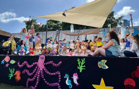 Palco com várias dezenas de crianças (e alguns adultos). Em primeiro plano, na parte frontal do palco, algumas imagens estilizadas com referências marítimas. Os rostos visíveis das crianças foram digitalmente desfocados.