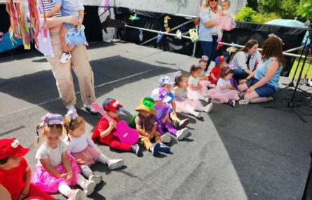 Palco com 13 crianças sentadas no piso do mesmo. Junto das crianças estão quatro pessoas adultas. Os rostos visíveis das crianças foram digitalmente desfocados.