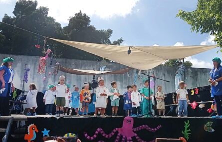 Palco com duas dezenas de crianças (e duas pessoas adultas). Em primeiro plano, na parte frontal do palco, algumas imagens estilizadas com referências marítimas. Os rostos visíveis das crianças foram digitalmente desfocados.