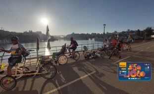 Grupo de 12 pessoas em bicicleta (normal ou adaptada) Fotografia junto do Rio Douro vendo-se do lado esquerdo parte da cidade de Gaia, à direita o Porto e, ao fundo, uma das pontes sobre o Rio Douro. No canto inferior esquerdo o logotipo da corrida.