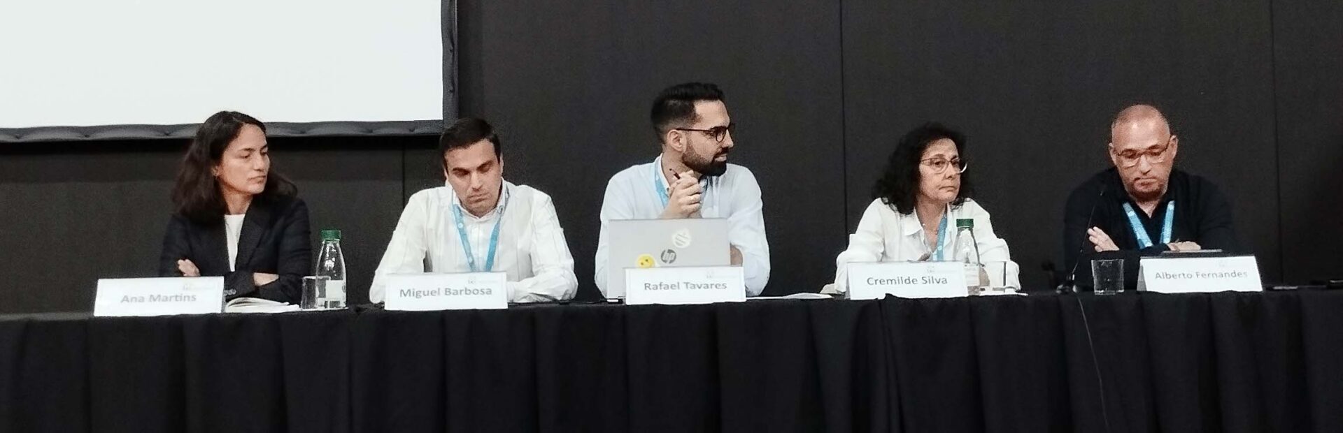 Cinco pessoas sentadas junto de uma mesa comprida (em palco). Na parede está uma tela (sem qualquer projeção).