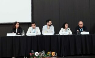 Cinco pessoas sentadas junto de uma mesa comprida (em palco). Na parede está uma tela (sem qualquer projeção).