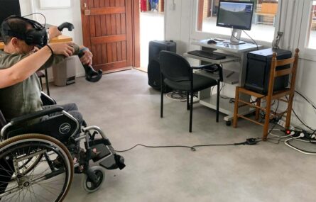 Uma pessoa, sentada em cadeira de rodas, utiliza diverso equipamento de realidade virtual. Junto dessa pessoa está uma mesa com diverso equipamento informático e um ecrã de computador.