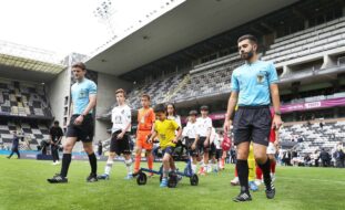 Vários jovens entram em campo relvado. São acompanhados por dois elementos da equipa de arbitragem e, ao centro, por uma criança que com um andarilho transporta uma bola de futebol. Ao fundo bancadas e público.