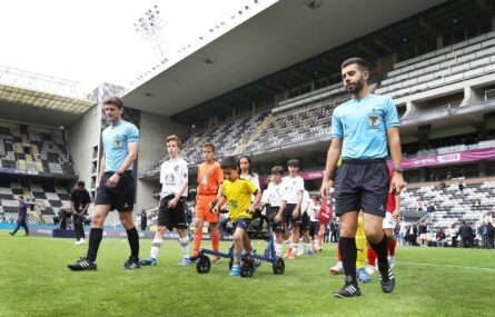 Vários jovens entram em campo relvado. São acompanhados por dois elementos da equipa de arbitragem e, ao centro, por uma criança que com um andarilho transporta uma bola de futebol. Ao fundo bancadas e público.