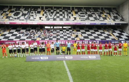 Duas equipas alinhadas no relvado de campo de futebol. Ao centro a dupla de arbitragem e um jovem com andarilho. Ao fundo bancadas e público.
