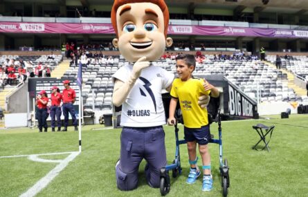 Uma criança (que se apoia em andarilho) ao lado da mascote chamada “O Ligas”. Estão no relvado de um estádio. Ao fundo da imagem bancadas e público.