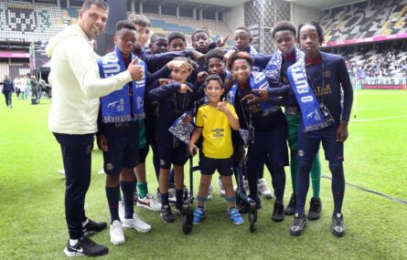 Grupo de 11 atletas e um adulto na companhia de uma criança (que se apoia em andarilho). Fotografia no relvado de estádio. Ao fundo bancadas e público.