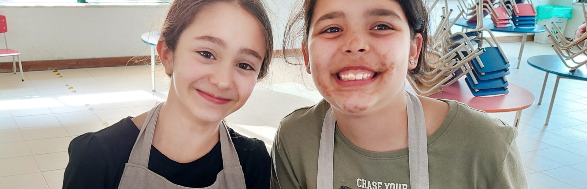 Duas crianças seguram em bacias azuis enquanto sorriem. Uma das crianças está com a cara lambuzada do que aparenta ser chocolate derretido. Mesas e cadeiras ao fundo da imagem.