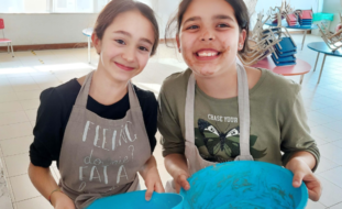 Duas crianças seguram em bacias azuis enquanto sorriem. Uma das crianças está com a cara lambuzada do que aparenta ser chocolate derretido. Mesas e cadeiras ao fundo da imagem.