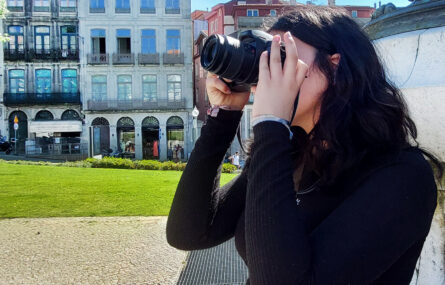 Jovem em praça pública utiliza máquina fotográfica para registar algo que não aparece na imagem. Relvado e edifícios antigos, ao fundo.