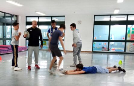 Cinco jovens e um adulto em atividade desportiva em ginásio interior. Um dos jovens está deitado no chão e tenta segurar uma bola.