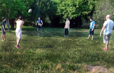 Cinco jovens em jardim relvado com árvores ao fundo. Aparentam estar a praticar atividade desportiva usando, para tal, uma bola. Uma pessoa adulta assiste.