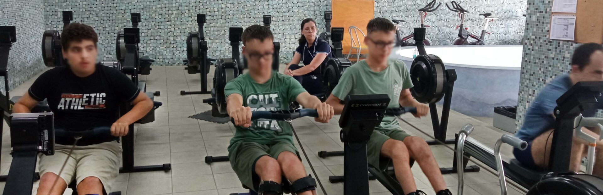 Quatro pessoas em espaço interior a utilizar equipamento de prática desportiva. Ao fundo, sentada, uma outra pessoa assiste. Os rostos visíveis dos quatro praticantes foram digitalmente desfocados.