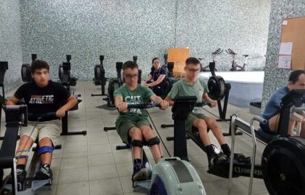 Quatro pessoas em espaço interior a utilizar equipamento de prática desportiva. Ao fundo, sentada, uma outra pessoa assiste. Os rostos visíveis dos quatro praticantes foram digitalmente desfocados.