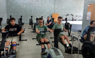 Quatro pessoas em espaço interior a utilizar equipamento de prática desportiva. Ao fundo, sentada, uma outra pessoa assiste. Os rostos visíveis dos quatro praticantes foram digitalmente desfocados.