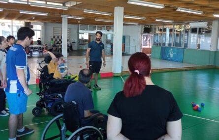Sete pessoas praticam boccia em espaço interior. Uma das pessoas prepara-se para lançar uma bola encarnada na direção de um conjunto de sete (azuis, encarnadas e branca) pousadas no chão.