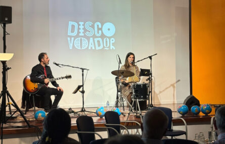 Duas pessoas no palco, uma delas com guitarra e a outra com um instrumento de percussão.