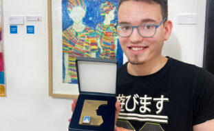 Fotografia de um jovem sorridente a segurar uma medalha nas mãos. Atrás dele, na parede, um quadro com uma pintura colorida.