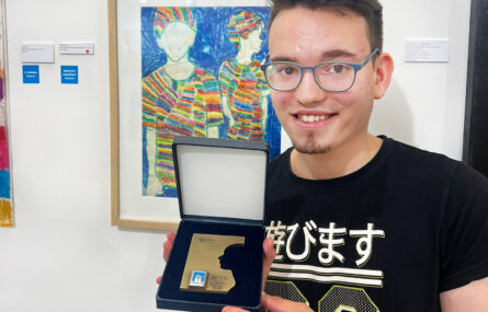Fotografia de um jovem sorridente a segurar uma medalha nas mãos. Atrás dele, na parede, um quadro com uma pintura colorida.