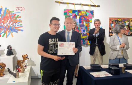 Fotografia de entrega de prémios e menções honrosas. Em destaque uma pessoa segura um diploma enquanto, ao lado, outra tem nas mãos uma medalha. Mesa com imensos diplomas e medalhas ainda por entregar e mais três pessoas ao fundo. Vários trabalhos expostos e pendurados na parede.