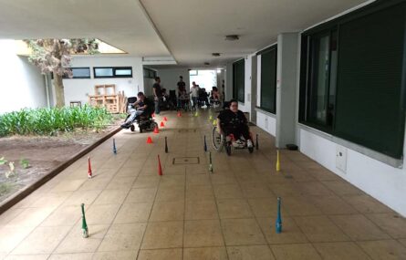 duas pessoas utilizadoras de cadeiras de rodas, participam em percurso de obstáculos. Ao fundo cerca de sete pessoas assistem.