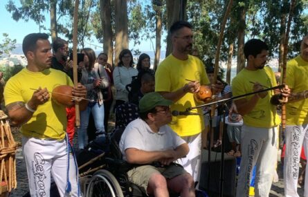 Grupo de pessoas num jardim. em primeiro plano quatro pessoas, com veste de capoeira e instrumentos musicais na mão. Uma pessoa junto a um microfone.