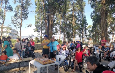Cerca de 50 pessoas num jardim com árvores altas e um bolo de aniversário colocado em cima da mesa.