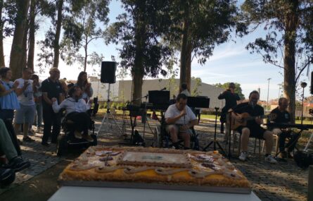Cerca de dez pessoas num jardim e um bolo de aniversário em cima de uma mesa. Uma pessoa em um microfone na mão. Uma segunda pessoa está a tocar guitarra e uma terceira piano.